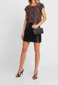 Blouse à motif floral dans des couleurs sombres, associée à une mini-jupe en cuir noir élégante avec une fente sur le côté. Accessoirisée avec un petit sac à main noir.