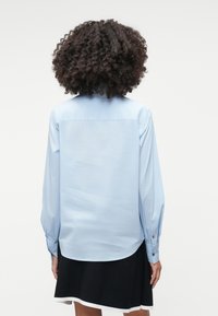 Camisa azul-claro de mangas longas e punhos abotoados, com um corte direito e uma textura suave, exibida de costas.