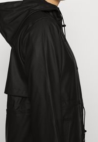 Chaqueta negra impermeable con capucha, cordón de ajuste y bolsillos frontales. Tejido suave con un acabado mate. Vista de perfil lateral.