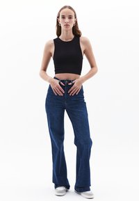 Top corto nero abbinato a jeans in denim blu a zampa d'elefante e sneakers grigie. I jeans hanno un alto punto vita e una chiusura a bottone visibile.