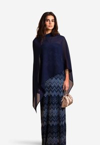 Poncho trasparente blu navy con scollo rotondo, sopra una gonna lunga a fantasia in tonalità di blu, abbinato a una piccola borsetta metallica.