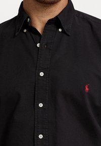 Svart skjorta med knappar i bomullstyg, med vita knappar och en liten röd broderad logotyp på vänster bröst.