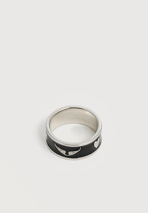 Bague argentée et noire avec des symboles gravés d’aile et de cœur sur la bande extérieure, présentée sur un fond gris clair uni.