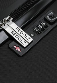 Gros plan sur une valise noire texturée avec des zips en argent portant l'inscription "REDOLZ" et un cadenas à combinaison TSA.