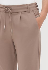 Pantalons beiges ajustés avec taille à cordon, design plissé, poches latérales et léger éclat dans un tissu doux et lisse.