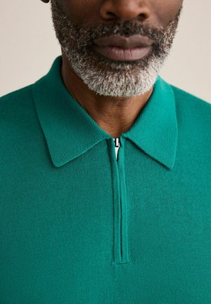 Közeli kép egy férfiról, akinek só-bors színű szakálla van, és türkizkék kötött galléros pólót visel, amelynek a gallérján rövid cipzár található.