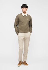 Νέος άνδρας με χακί πράσινο πουλόβερ πάνω από λευκό πουκάμισο με γιακά, μπεζ παντελόνι και καφέ σουέντ αθλητικά παπούτσια, όρθιος με τα χέρια στις τσέπες.