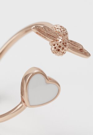 Åben ring i rosaguld med et bi-design i den ene ende og en hvid hjerteform i den anden ende mod en lys baggrund.