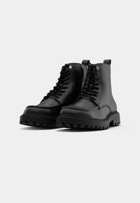 Paire de bottines noires en cuir à lacets avec semelles épaisses et robustes et fermeture éclair latérale sur fond uni clair.