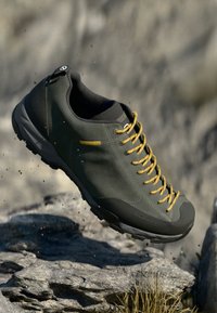 Dunkelgrüner Wanderschuh mit gelben Schnürsenkeln, strukturiertem Obermaterial und verstärkter Zehenschutzkappe. Ideal für unwegsames Gelände mit griffiger Sohle.