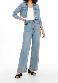 Mujer con chaqueta de mezclilla recortada azul claro y jeans de pierna ancha de cintura alta, sandalias de tacón negro y camiseta blanca debajo.