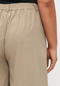 Gros plan d'une main d'une personne reposant à côté d'un pantalon beige à taille élastique et d'un haut noir, montrant la vue arrière et latérale.