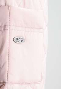 Helle rosa gesteppte Jackentasche mit sichtbaren Nähten und einem kleinen ovalen Metallabzeichen mit der Aufschrift "edc SINCE 980."