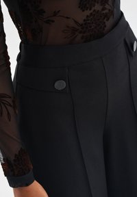 Czarne spodnie z wysokim stanem o gładkiej teksturze, z dwoma guzikami z przodu i wyraźnymi fałdami dla nadającego struktury wyglądu.