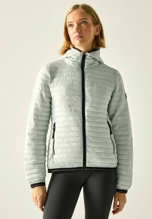 Donna che indossa una giacca trapuntata grigio chiaro con cappuccio e zip nera, abbinata a leggings neri, in piedi davanti a uno sfondo semplice.
