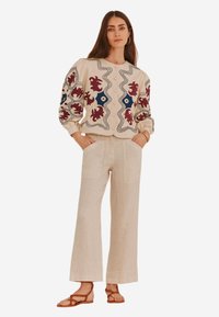 Blusa beige ricamata con motivi floreali e bordi scallop, abbinata a pantaloni larghi in lino chiaro e sandali minimalistici marroni.