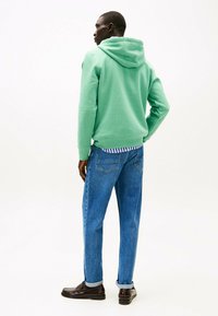 Sudadera verde con capucha y un corte relajado, combinada con jeans de mezclilla azul claro. Unos zapatos marrones completan el atuendo casual.