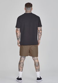 T-shirt noir à manches courtes, shorts marron, tatouages visibles, et baskets noires avec semelles blanches, se tenant devant un fond clair.