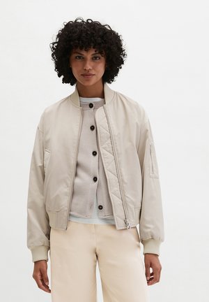 Woman with curly hair wearing a light beige bomber jacket over a cream buttoned cardigan and matching light beige pants against white background.