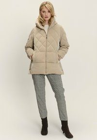 Giacca imbottita beige con un motivo a rombi, chiusura con zip e cappuccio integrato. Il modello indossa pantaloni a quadri grigi e stivali alla caviglia marroni.