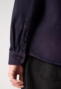 Cămașă din denim închis, cu mâneci lungi, având butoane negre și cusături contrastante de culoare purpurie. O mână este vizibilă, ținând țesătura aproape de talie.