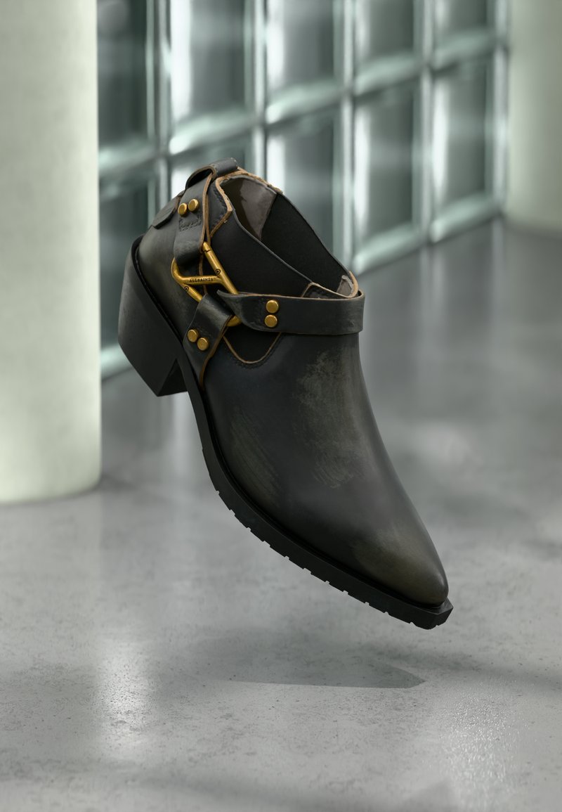 Μαύρο δερμάτινο παπούτσι με τακούνι, με χρυσή πόρπη και λουράκια, τοποθετημένο σε αντανάκλαση τσιμεντένιου δαπέδου, με φόντο τοίχο από γυάλινα τουβλάκια.