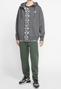 Grå zip-up hoodie med framfickor och dragsnoddar, mönstrad svart och vit skjorta under, gröna byxor och svarta sneakers med grå accenter.