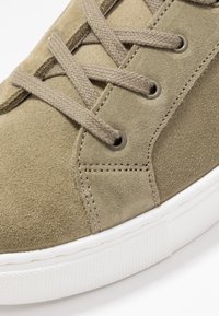 Gros plan d'une basket en daim beige avec des lacets tissés passant dans des œillets métalliques et une semelle en caoutchouc blanche texturée.