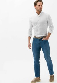 Camicia bianca con bottoni, jeans blu, cintura intrecciata marrone e scarpe beige slip-on. Fit slim con una texture liscia e design casual.