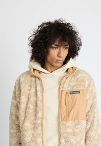 Jachetă din fleece pufoasă bej, cu un buzunar din nylon bej și un logo pe piept. Are glugă și o textură netedă.