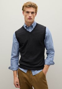 Junger Mann mit blondem Haar, der ein blaues Hemd unter einem schwarzen ärmellosen Pullover trägt und braune Hosen anhat, steht vor einer hellen Wand.