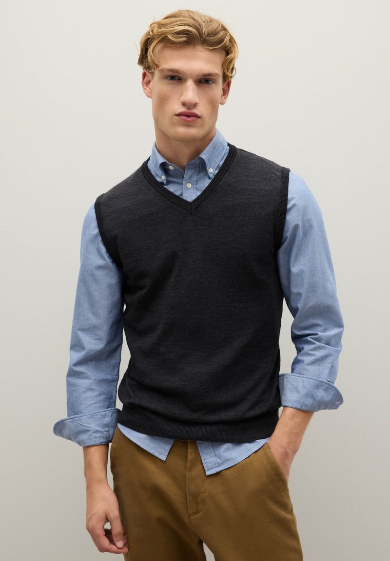 Junger Mann mit blondem Haar, der ein blaues Hemd unter einem schwarzen ärmellosen Pullover trägt und braune Hosen anhat, steht vor einer hellen Wand.