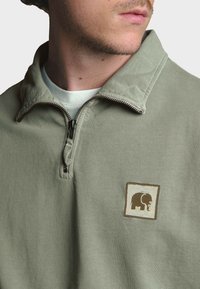Sudadera verde claro tipo quarter-zip con una textura suave, presentando un logo de elefante en un parche cuadrado en el lado izquierdo del pecho. Cierre con cremallera metálica.