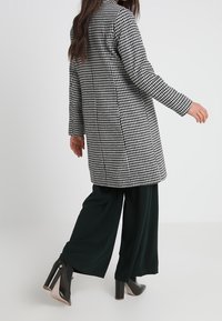 Schwarzer und weißer Hahnentrittmuster-Mantel mit langen Ärmeln, geradem Schnitt und Kragen, kombiniert mit dunklen weit geschnittenen Hosen und schwarzen Absatzstiefeln.