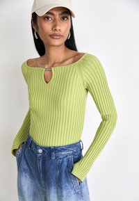 Top a coste verde con dettagli a chiave sul davanti, abbinato a pantaloncini di jeans azzurri a vita alta. Design semplice con maniche lunghe.