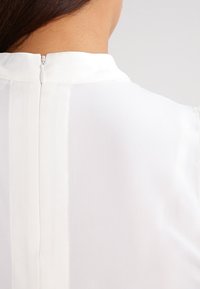 Blusa blanca con cuello alto, que presenta una cremallera oculta en la parte trasera y una tela suave y texturizada. Incluye detalles de costura vertical.