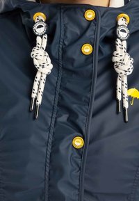 Veste imperméable navy avec fermeture éclair, deux boutons jaunes et cordons de serrage blancs avec embouts et motifs noirs.