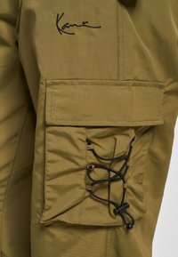 Poche de pantalon cargo marron avec un cordon noir réglable et une signature noire cousue au-dessus de la poche.