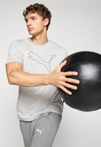 Grå t-shirt med korte ærmer og sort Puma-logo; lysegrå sweatpants med hvidt Puma-logo; holder en sort medicinbold.