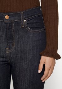 Jean en denim foncé avec une coupe ajustée, coutures visibles en orange, et un design classique à cinq poches. Manchon de pull côtelé marron qui dépasse.