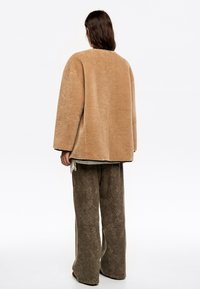 Chaqueta de forro polar oversize color beige con una textura suave, cuello redondo y hombros caídos, combinada con pantalones anchos marrones de estampado.