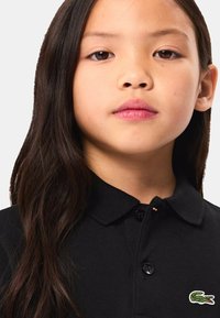 Czarna koszulka polo z prążkowanym kołnierzykiem i trzema guzikami, z zielonym logo krokodyla po lewej stronie. Gładki materiał, dopasowany krój.