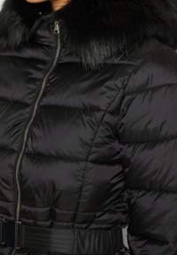 Kobieta ubrana w czarną pikowaną zimową kurtkę z zamkiem z przodu, paskiem w talii i czarnym kołnierzem wykończonym futrem.