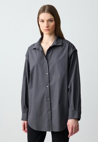 Chemise grise à manches longues avec col, au fit décontracté. Le tissu semble lisse avec un léger éclat. Fermeture à boutons à l'avant.