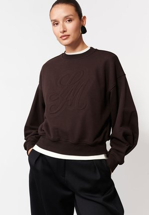 Kobieta ubrana w ciemno-brązowy, oversize'owy sweter z haftowanymi inicjałami, czarne spodnie w szerokie nogawki oraz duże czarno-złote kolczyki.