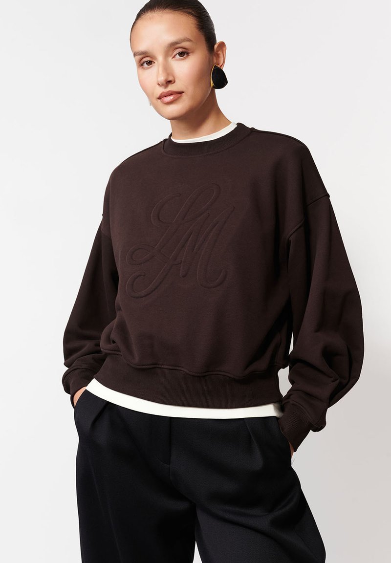 Kobieta ubrana w ciemno-brązowy, oversize'owy sweter z haftowanymi inicjałami, czarne spodnie w szerokie nogawki oraz duże czarno-złote kolczyki.