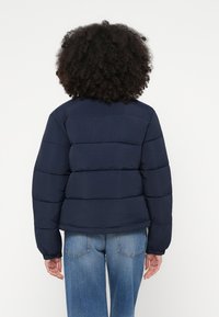 Tumši zilā pūkaina jaka ar horizontālu nodalījumu dizainu, elastīgām aprocēm un apaļu kakla izgriezumu. Valkāta kopā ar gaiši zilām taisna silueta džinsiem.