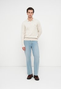 Strickpullover in Creme mit Zopfmuster und Polokragen, kombiniert mit hellblauen geraden Jeans und braunen Schuhen, vor einem weißen Hintergrund.