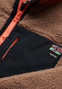 Fleece jacket featuring a tan body, navy fabric panel, coral zipper, and a multicolored label. Textured and warm design.