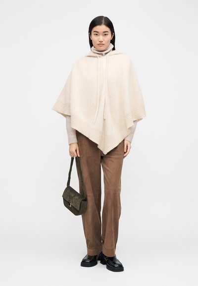Poncho beige clair avec capuche, bordures côtelées et cordons de serrage. Associé à un pull gris clair et un pantalon en velours côtelé marron, tenant un sac vert.